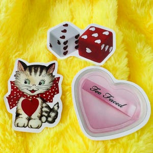 May include: Three stickers featuring a white die with red hearts, a red die with white hearts, a kitten with a red polka dot bow and a heart on its chest, and a heart-shaped pink compact with the text Too Faced printed on the surface.
