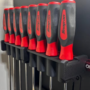 May include: A collection of ten screwdrivers with black and red handles, arranged in a black holder. The handles display the "Snap-on" logo. A red metal bar with the text "Olsatools" is attached to the holder.