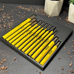 Op de afbeelding: Een set gele en zwarte beitels in een zwarte lade. De beitels zijn in een rij gerangschikt, met de gele handvatten naar de kijker gericht. De lade staat op een donkere ondergrond met houtkrullen en twee potplanten.