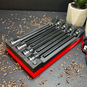 May include: A set of black and silver hex keys in a red and black organiser. The organiser is on a dark surface with scattered wood shavings. A chrome ratchet wrench is next to the organiser. Two small potted plants are in the background.