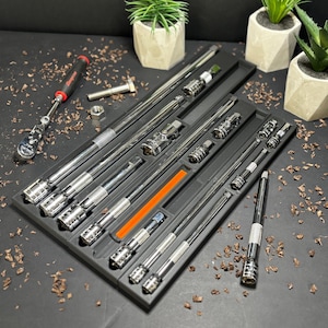 Może przedstawiać: Zestaw chromowanych nasadek i przedłużeń ułożonych w czarnej tacy. Widoczny jest również czerwono-czarny klucz zapadkowy, nakrętka i śruba. Tło obejmuje małe rośliny doniczkowe i dekoracyjne wióry drewniane.