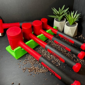May include: A collection of red and black hammers with the word "ICON" printed on the handles. The hammers are arranged on a green stand, with small potted plants in the background. The hammers have a red head and a black handle.