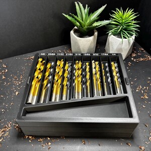 May include: A set of gold and silver drill bits in a black plastic organiser. The drill bits are arranged by size, with measurements in inches printed on the organiser. Two small potted succulents are in the background.