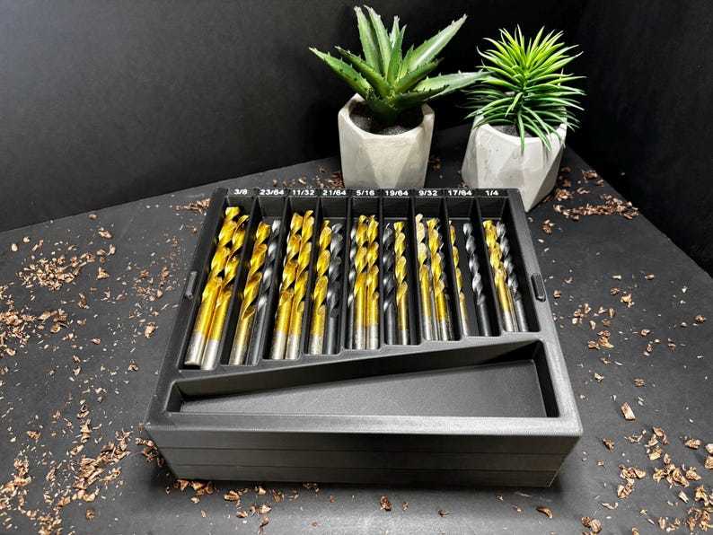 May include: A set of gold and silver drill bits in a black plastic organiser. The drill bits are arranged by size, with measurements in inches printed on the organiser. Two small potted succulents are in the background.