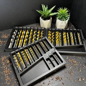 Może przedstawiać: Zestaw wierteł w czarnym organizerze. Wiertła mają złote i srebrne wykończenie. Dwie małe rośliny doniczkowe znajdują się w tle. Wiertła są ułożone według rozmiaru.