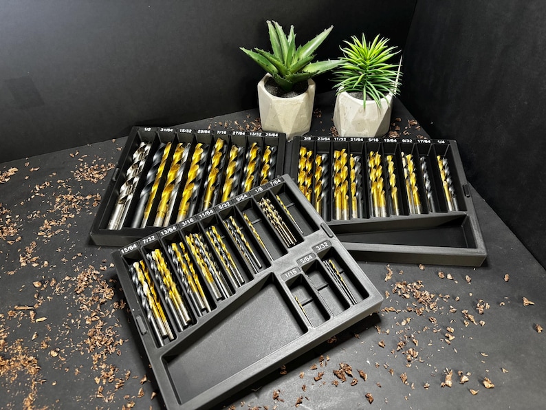 May include: A set of drill bits in a black organiser. The drill bits have a gold and silver finish. The organiser is on a black surface with two small potted plants in the background.