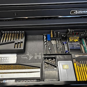 May include: A black Husky tool chest drawer filled with various drill bits, drivers, and other tools. The tools are organised in trays and cases, with gold and silver drill bits. The drawer is open, revealing the contents.