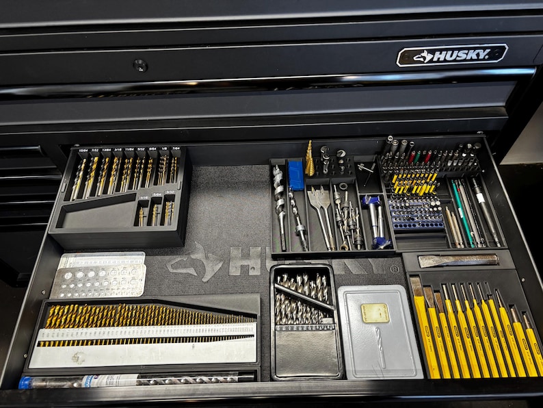 May include: A black Husky tool chest drawer filled with various drill bits, drivers, and other tools. The tools are organised in trays and cases, with gold and silver drill bits. The drawer is open, revealing the contents.