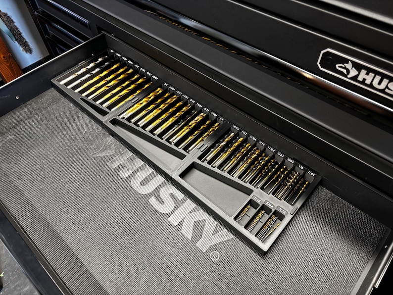 May include: A black tool chest drawer containing a selection of drill bits. The bits are arranged in a black tray, with sizes marked. The drill bits have a gold and silver colour scheme.