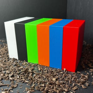 May include: Six rectangular blocks in white, black, green, orange, blue, and red are arranged in a row. They are placed on a surface covered in wood shavings. Each block is the same size and shape.