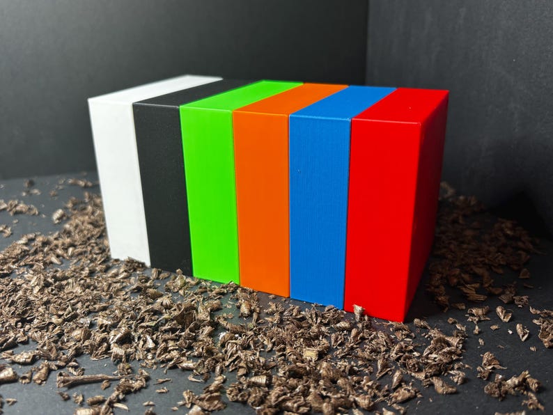 May include: Six rectangular blocks in white, black, green, orange, blue, and red are arranged in a row. They are placed on a surface covered in wood shavings. Each block is the same size and shape.