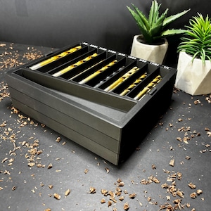 May include: A black drill bit organiser with multiple compartments, each holding gold and silver drill bits. The organiser sits on a dark surface with wood shavings, and two small potted plants are in the background.