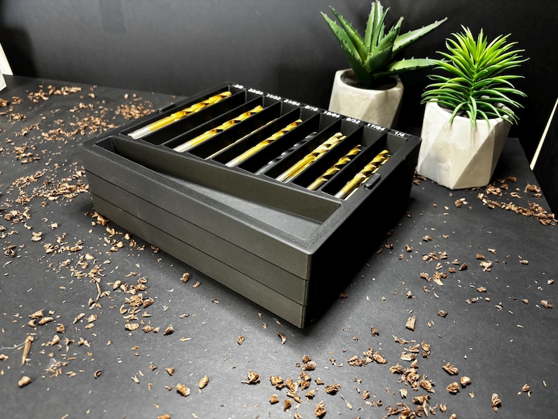 May include: A black drill bit organiser with multiple compartments, each holding gold and silver drill bits. The organiser sits on a dark surface with wood shavings, and two small potted plants are in the background.