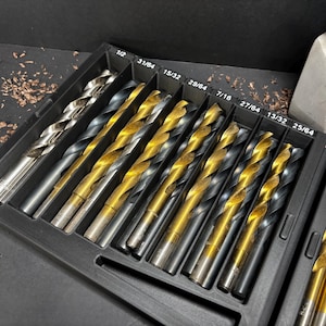 May include: A set of drill bits in a black plastic case. The drill bits have silver and gold finishes and are in various sizes. The case is labelled with the sizes of the drill bits. A small plant in a white pot is in the background.