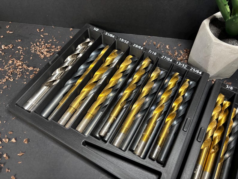 May include: A set of drill bits in a black plastic case. The drill bits have silver and gold finishes and are in various sizes. The case is labelled with the sizes of the drill bits. A small plant in a white pot is in the background.