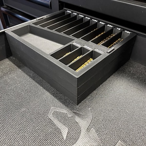 May include: A black drill bit organiser with labelled compartments, holding various sizes of gold-coloured drill bits. The organiser is inside a drawer, with the sizes printed in white. The drawer is in a black cabinet.