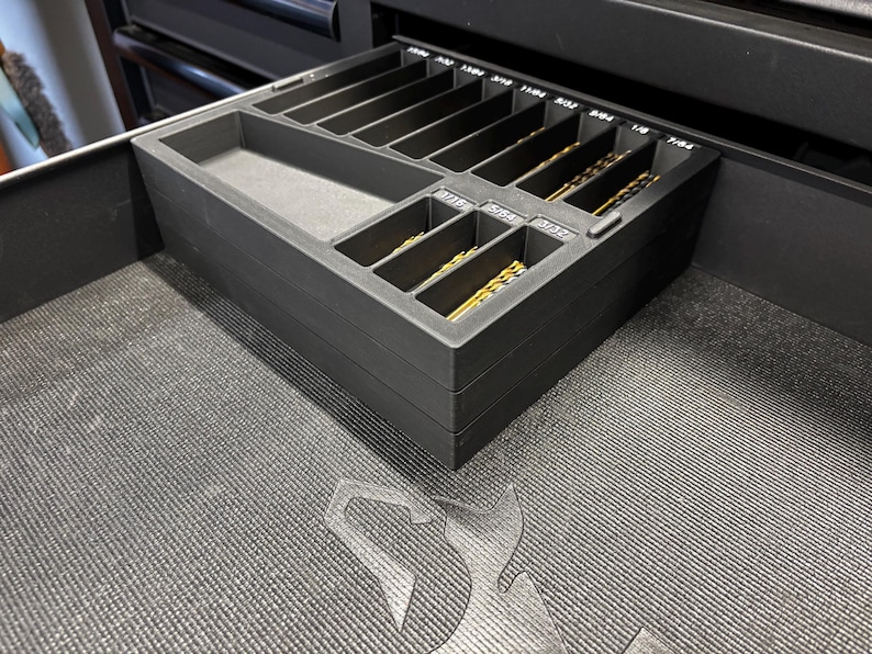 May include: A black drill bit organiser with labelled compartments, holding various sizes of gold-coloured drill bits. The organiser is inside a drawer, with the sizes printed in white. The drawer is in a black cabinet.