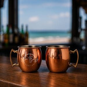 May include: Two copper-colored Moscow Mule mugs with gold-colored handles sit on a wooden bar. The mugs have a shiny finish and a logo on the front. The background shows a beach scene with the ocean and sky visible through a window.