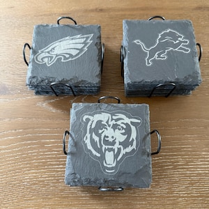 May include: Three stacks of square dark gray slate coasters held in black metal wire holders. Each stack features a different engraved sports team logo: an eagle, a lion, and a bear. The coasters have rough, natural edges and a flat surface.