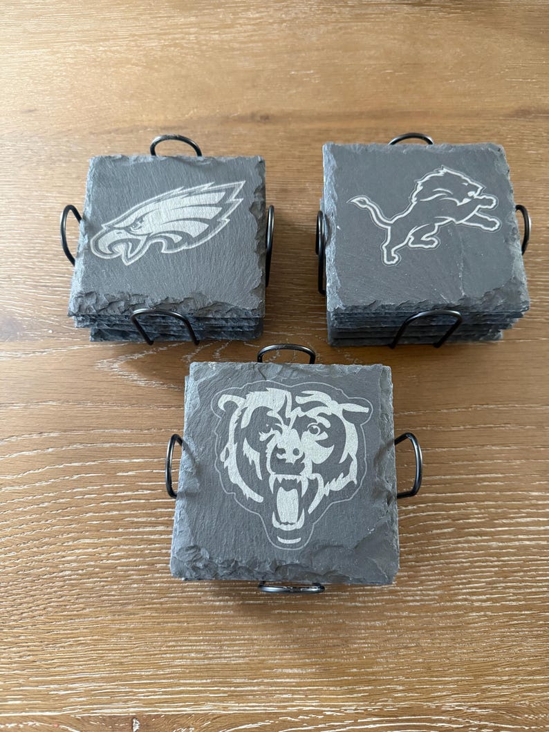 May include: Three stacks of square dark gray slate coasters held in black metal wire holders. Each stack features a different engraved sports team logo: an eagle, a lion, and a bear. The coasters have rough, natural edges and a flat surface.