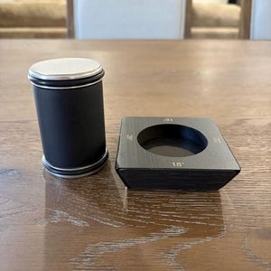 May include: A matte black cylindrical tool with a silver top and bottom rim, positioned next to a square black base featuring a circular cutout. The base has the numbers 18, 20, and 22 engraved on its edges, indicating specific size measurements.