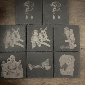 May include: Eight square dark gray slate coasters featuring laser-engraved images. Two show a dog, two show a baby with a dog wearing reindeer antlers, one shows a person holding a baby, one shows a baby in a seat, one shows a person reclining, and one shows a baby with the name Everett.