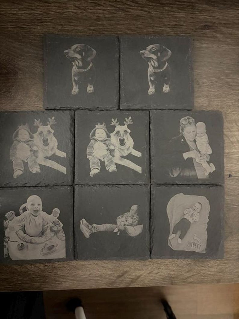 May include: Eight square dark gray slate coasters featuring laser-engraved images. Two show a dog, two show a baby with a dog wearing reindeer antlers, one shows a person holding a baby, one shows a baby in a seat, one shows a person reclining, and one shows a baby with the name Everett.