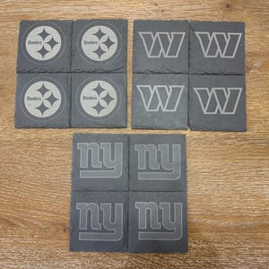 May include: A set of twelve square slate coasters featuring engraved sports team logos. Four coasters display the Pittsburgh Steelers logo, four feature the Washington Commanders W logo, and four show the New York Giants ny logo in gray.