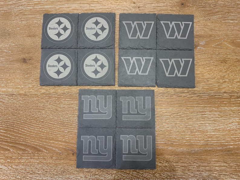 May include: A set of twelve square slate coasters featuring engraved sports team logos. Four coasters display the Pittsburgh Steelers logo, four feature the Washington Commanders W logo, and four show the New York Giants ny logo in gray.