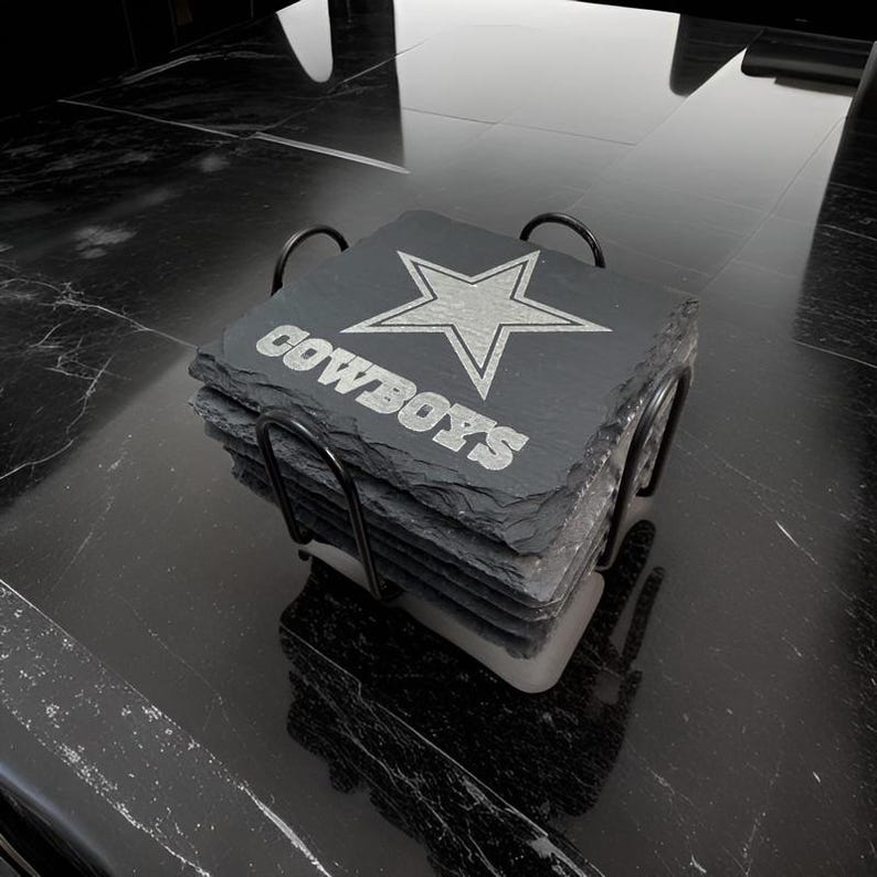 May include: A stack of four square black slate coasters held in a black metal wire rack. Each coaster features an engraved silver-toned star emblem and the text COWBOYS in a bold, block font. The coasters have natural, rough-hewn edges.