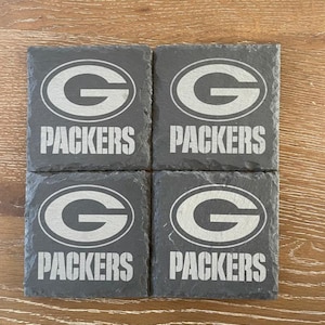 May include: A set of four square dark gray slate coasters, each featuring the Green Bay Packers logo and the word PACKERS engraved in a light gray tone. The coasters have a natural, rough-hewn edge texture and are arranged in a two-by-two grid.