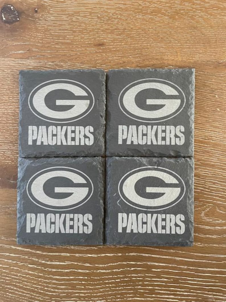 May include: A set of four square dark gray slate coasters, each featuring the Green Bay Packers logo and the word PACKERS engraved in a light gray tone. The coasters have a natural, rough-hewn edge texture and are arranged in a two-by-two grid.