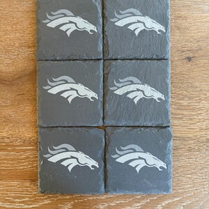 May include: A set of six square dark gray slate coasters, each featuring a light gray engraved graphic of a stylized horse head profile with a flowing mane, arranged in two columns of three on a light brown wood grain surface.