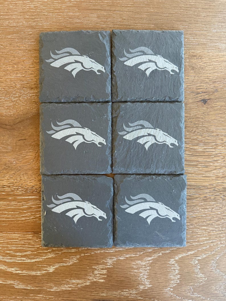 May include: A set of six square dark gray slate coasters, each featuring a light gray engraved graphic of a stylized horse head profile with a flowing mane, arranged in two columns of three on a light brown wood grain surface.