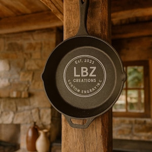 May include: A black cast iron skillet with a long handle is mounted on a wooden beam. The skillet has the text "Est. 2023 LBZ CREATIONS CUSTOM ENGRAVING" in a circular design. The background includes a stone wall and a window.
