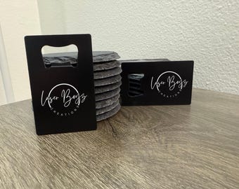 May include: Two black rectangular metal bottle openers featuring the white text Laser Boyz Creations in a circular logo design, positioned in front of a stack of round dark gray slate coasters with rough, textured edges.