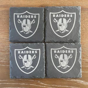 May include: A set of four square dark gray slate coasters, each featuring the engraved Raiders logo with a football helmet and crossed swords. The text RAIDERS is clearly visible at the top of each shield-shaped emblem on the textured stone surface.