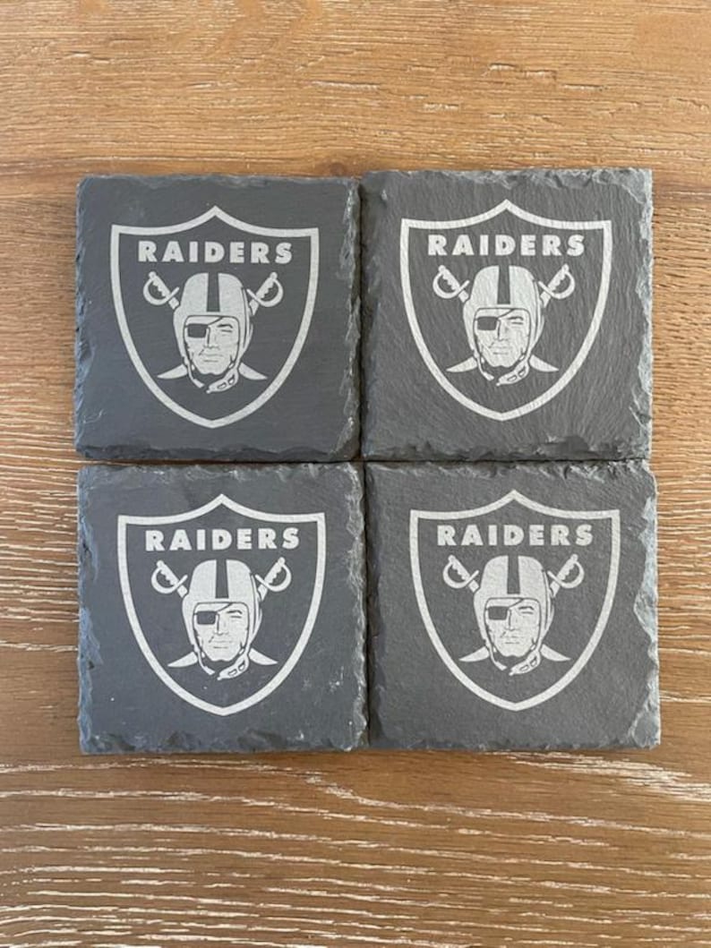 May include: A set of four square dark gray slate coasters, each featuring the engraved Raiders logo with a football helmet and crossed swords. The text RAIDERS is clearly visible at the top of each shield-shaped emblem on the textured stone surface.