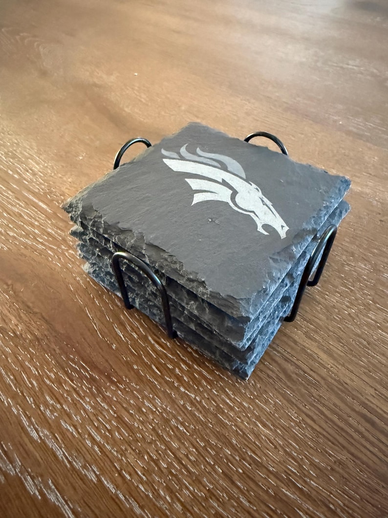 May include: A stack of four square dark gray slate coasters with rough, natural edges held in a black metal wire holder. Each coaster features a white engraved graphic of a stylized horse head profile with a flowing mane facing to the right.