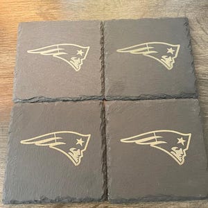 May include: A set of four square dark gray slate coasters with rough, natural edges. Each coaster features a light-colored, engraved New England Patriots logo, depicting a stylized profile of a person wearing a tricorne hat with a star.