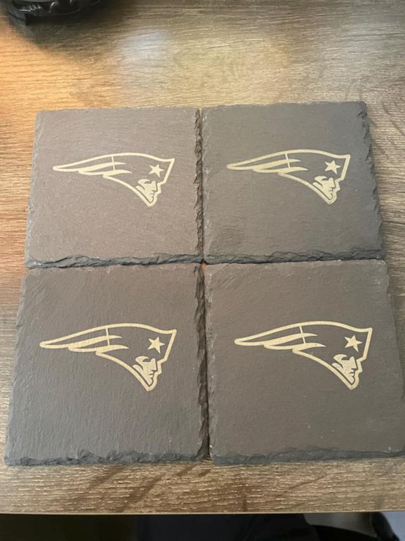 May include: A set of four square dark gray slate coasters with rough, natural edges. Each coaster features a light-colored, engraved New England Patriots logo, depicting a stylized profile of a person wearing a tricorne hat with a star.