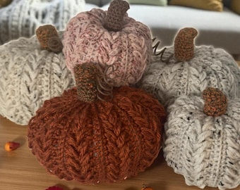May include: A collection of handmade knitted pumpkins in various colors, including orange, pink, and gray. Each pumpkin has a brown stem and a textured, cable-knit design. The pumpkins are arranged on a wooden surface, with small decorative gourds and fall foliage.
