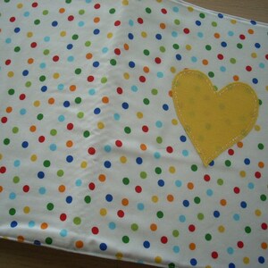 May include: A white fabric with a yellow heart-shaped patch sewn on. The fabric has a polka dot pattern with red, blue, green, and yellow dots.