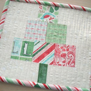 以下が含まれることがあります： 赤、緑、白のストライプの縁取りが施された白いキルティング生地の正方形。正方形の中央は、緑と白の格子柄、赤と白のストライプ柄、花柄など、さまざまなパターンでパッチワークされた正方形で構成されています。左下の角には「LL」の文字が見えます。