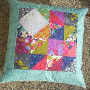 May include: A turquoise pillow with a patchwork design featuring various colors and patterns, including pink, purple, red, green, and blue. The pillow has a white border and a white star pattern on the back.