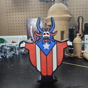 May include: A 3D printed figurine of a cartoon monster wearing a Puerto Rican flag. The monster has blue skin, red horns, and a white star on its chest. The figurine is standing on a black base.