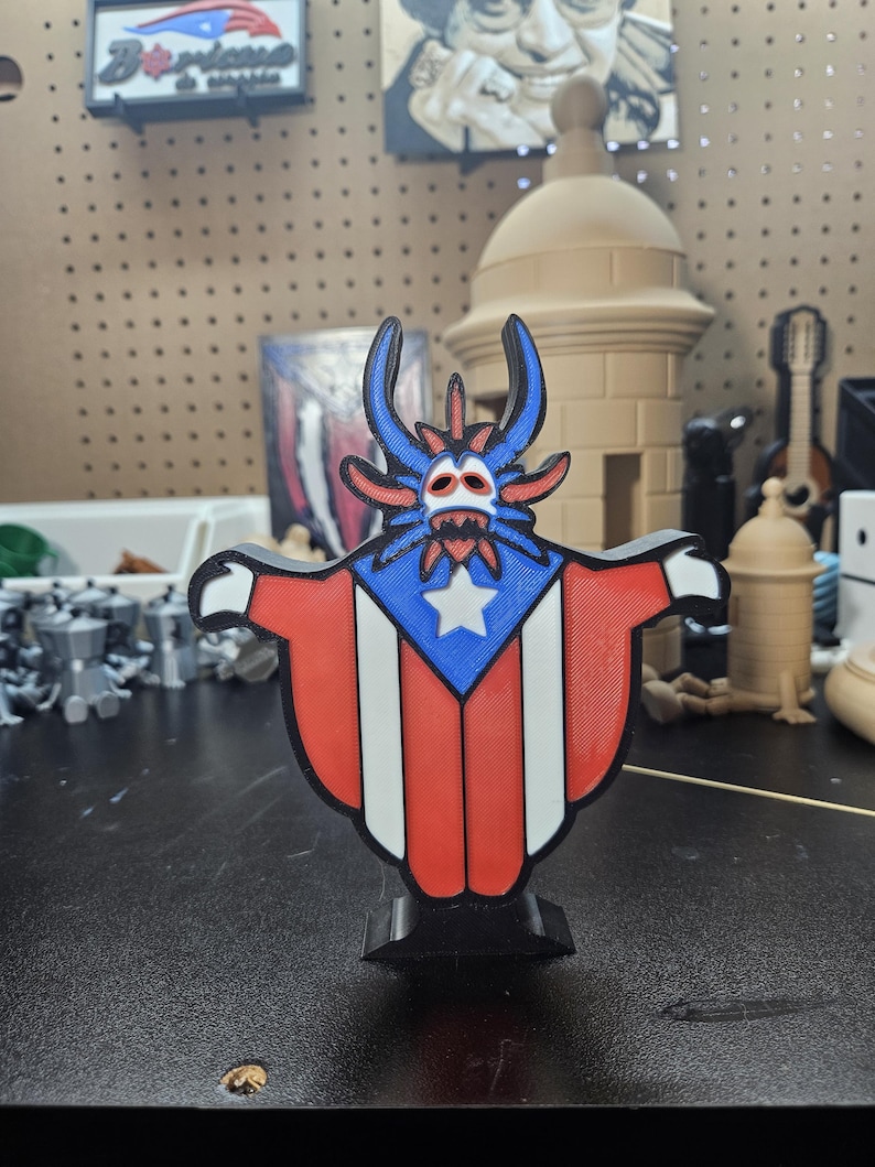 May include: A 3D printed figurine of a cartoon monster wearing a Puerto Rican flag. The monster has blue skin, red horns, and a white star on its chest. The figurine is standing on a black base.
