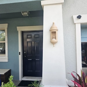 May include: Exterior view of a house entrance with a dark blue door and a decorative beige wall-mounted lantern. The house has a light blue and white color scheme, with green plants in the foreground.