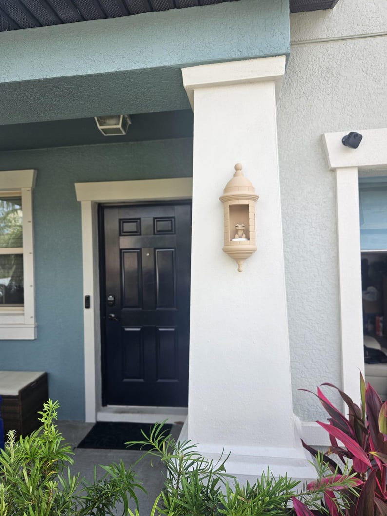 May include: Exterior view of a house entrance with a dark blue door and a decorative beige wall-mounted lantern. The house has a light blue and white color scheme, with green plants in the foreground.