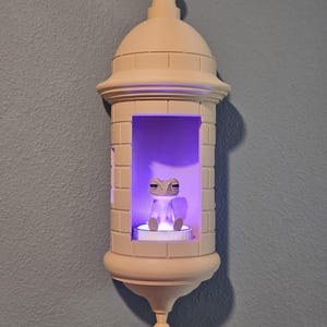 May include: A beige, castle-shaped wall sconce with a small frog figurine inside, illuminated by a purple light. The sconce has a domed top and a decorative base, mounted on a blue wall.